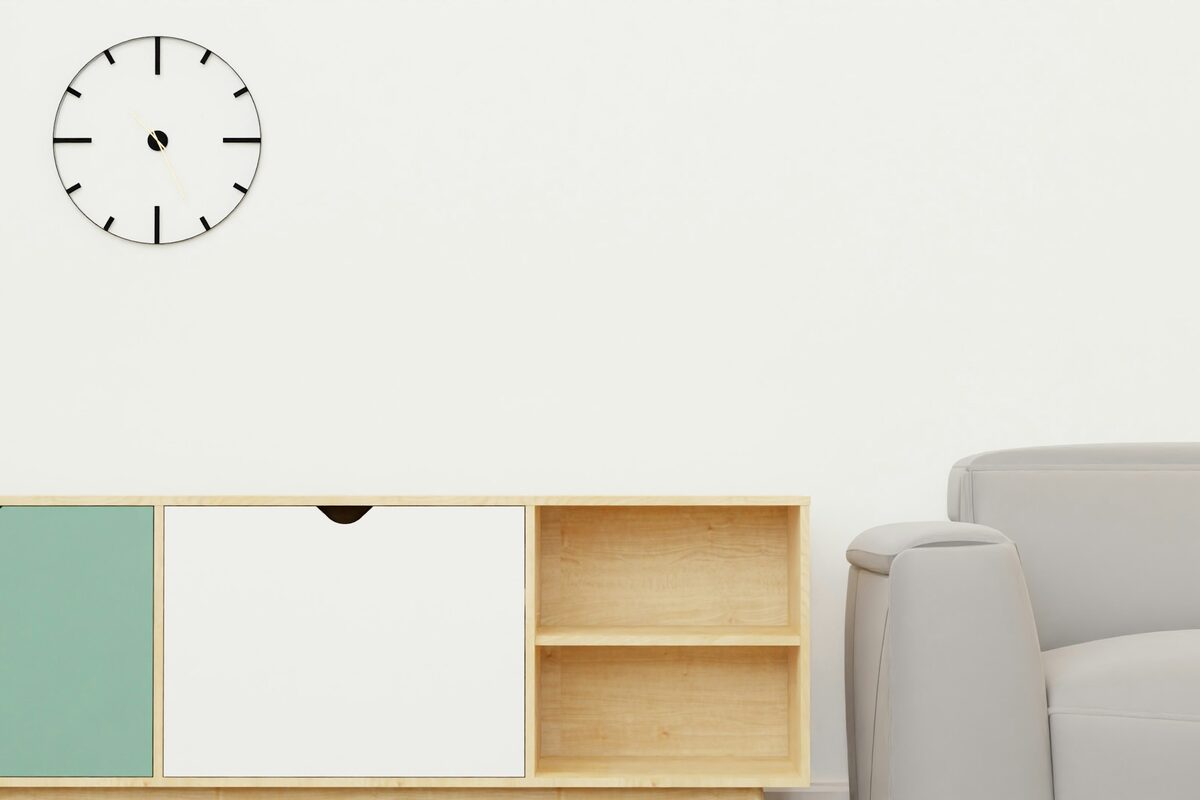 Horloge ronde à index noirs sur un mur blanc, au-dessus d'un meuble bas en bois clair et d'un fauteuil gris